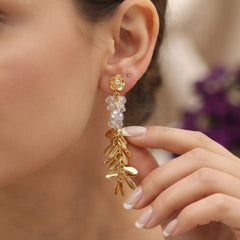 A close-up of a woman wearing a dangling gold earring with a flower-shaped stud and leaf-like drops, alongside a hand with manicured nails.