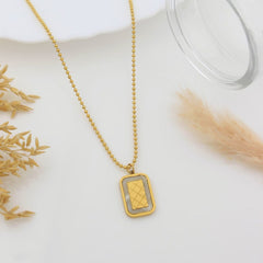 A gold ball-chain necklace with a rectangular pendant featuring a textured gold grid pattern on a silver-toned background, styled on a white surface with dried grass and a glass plate.