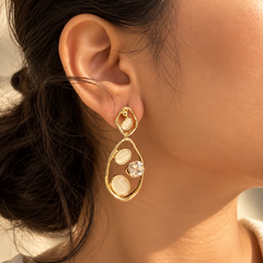 A close-up of a woman's ear wearing a gold teardrop-shaped dangle earring with cream-colored oval stones and a clear faceted crystal accent.