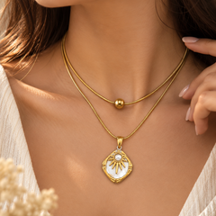 A close-up lifestyle shot of a model wearing a double-layered gold necklace. The top layer is a delicate chain with a small gold bead, and the bottom layer features a gold pendant with a white inlay and a sunburst design.