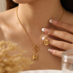A close-up lifestyle shot of a woman wearing a delicate gold chain necklace with a Y-shaped design. The necklace features a small square pendant engraved with the letter 'H', a larger padlock-shaped pendant with an 'H' cutout, and a toggle clasp.