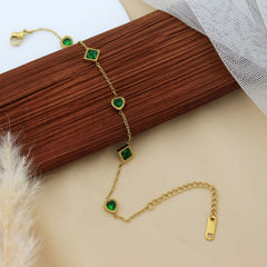 A gold chain bracelet with five green gemstone accents, styled on a brown wooden block with beige fabric and dried grass in the background.