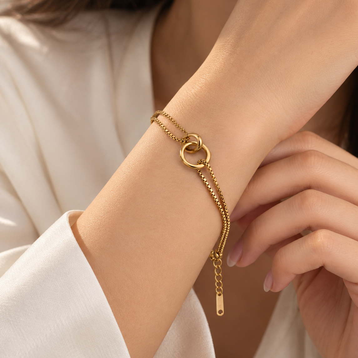 A delicate gold chain bracelet with a double interlocking ring closure and a dangling chain extender, shown worn on a woman's wrist.