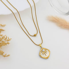 A gold-toned necklace with a textured, irregularly shaped pendant featuring a sunburst design and a central white stone, displayed on a white surface with dried botanical props.