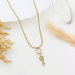 A gold chain necklace with a dangle pendant featuring a white moonstone and clear rhinestones, styled on a white surface with dried grass and a glass rim.