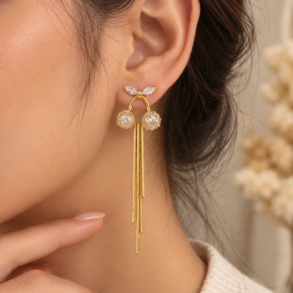 A close-up of a woman's ear wearing a gold dangle earring with clear crystal-like stones and three hanging gold strands.