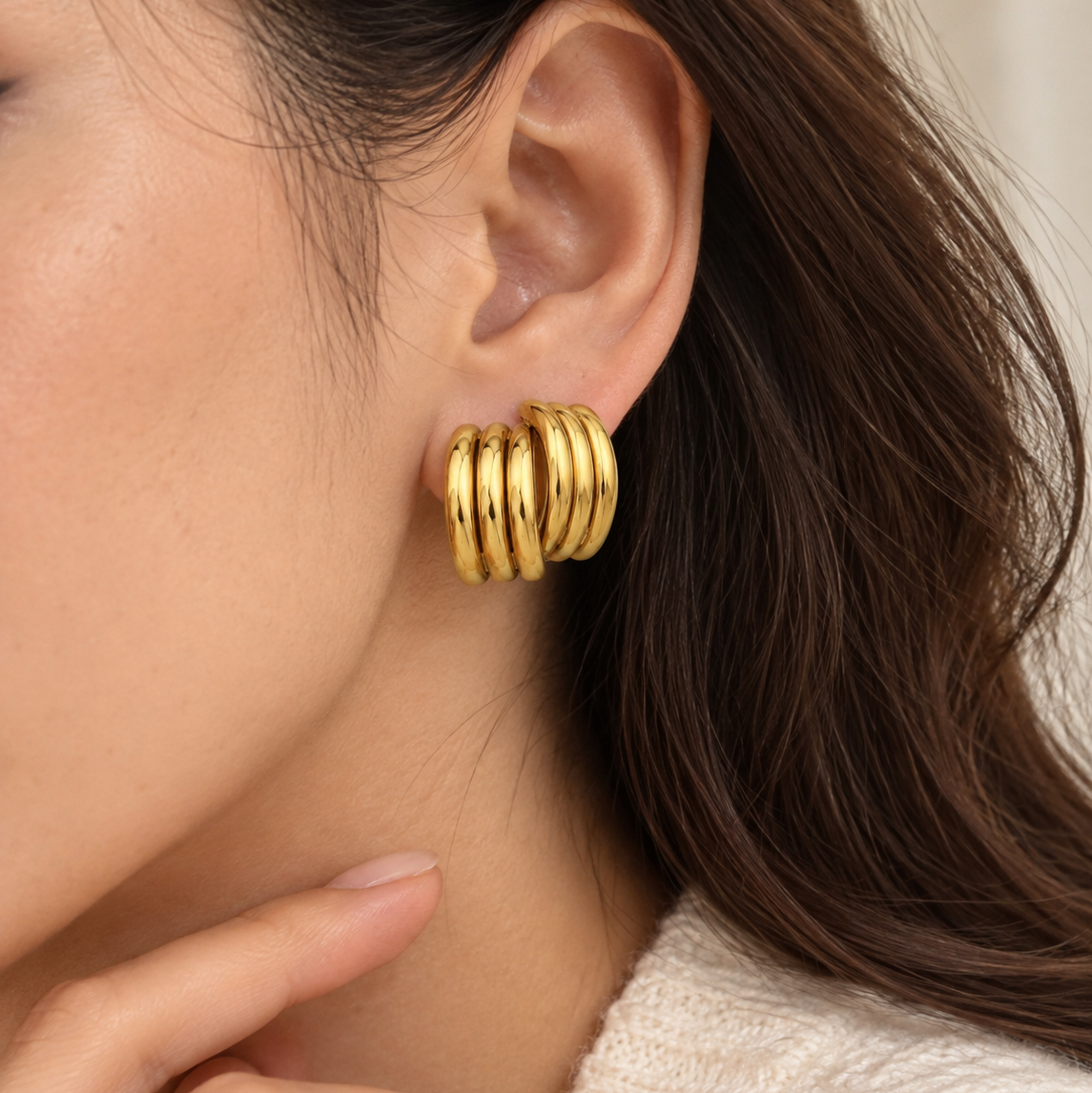 A close-up of a model's ear wearing a stack of three polished gold hoop earrings, with her hand near her chin and a soft neutral background.