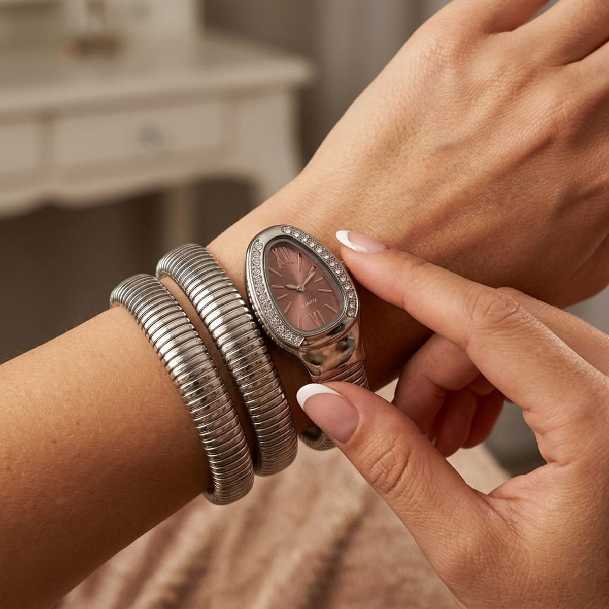 A close-up lifestyle shot of a woman's wrist wearing a silver-toned snake-style bangle bracelet and a silver-toned oval watch with a rose gold dial.