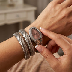 A close-up lifestyle shot of a woman's wrist wearing a silver-toned snake-style bangle bracelet and a silver-toned oval watch with a rose gold dial.