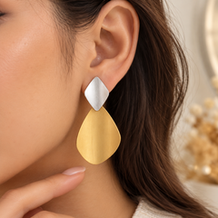 A close-up of a woman wearing a large statement earring with a silver diamond-shaped top and a gold teardrop-shaped drop, shown against her ear and neck.