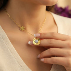 A close-up lifestyle shot of a woman wearing a delicate gold chain necklace with two small flower charms, one of which is a daisy pendant featuring a clear crystal center. She is wearing a cream-colored knit top, and her hand gently touches the pendant.