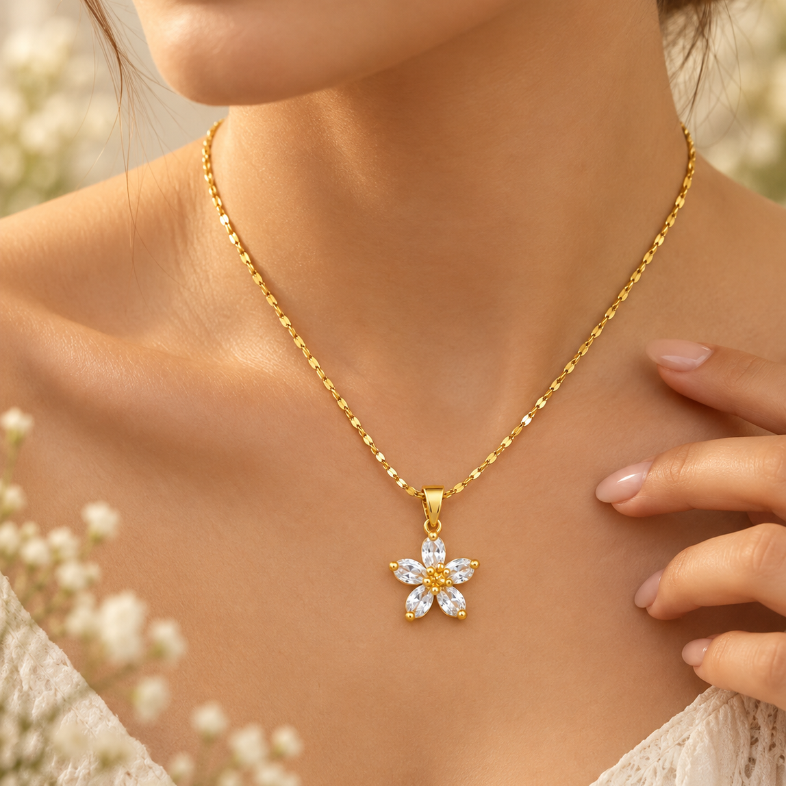 A close-up lifestyle shot of a woman wearing a delicate gold chain necklace with a flower-shaped pendant set with clear crystals, framed by soft white flowers and a warm, sunny background.