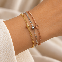 Three delicate chain bracelets in yellow gold, silver, and rose gold, each featuring a small polished bead, shown stacked on a person's wrist.