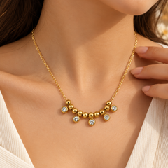 A close-up of a model wearing a delicate gold necklace with a curved row of small gold beads and five clear crystal-like round accents, paired with a white top.