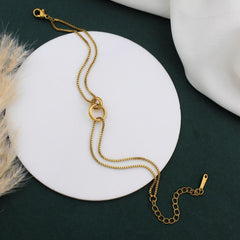 A gold-toned chain necklace with a double-ring pendant, displayed on a round white plate over a dark green background with soft decorative props.