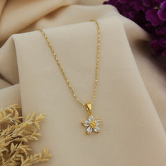 A gold chain necklace with a flower-shaped pendant featuring clear rhinestones, displayed on a beige fabric stand with dried flowers and purple flowers in the background.