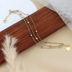 A delicate layered necklace with three chains in gold, silver, and rose gold, featuring small metallic beads and a gold heart charm, displayed on a wooden block with neutral background props.