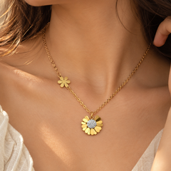 A close-up lifestyle shot of a model wearing a delicate gold chain necklace with two small flower charms and a larger flower pendant featuring a sparkling clear center.