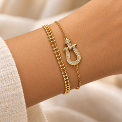 A close-up of a person's wrist wearing two gold bracelets: one chunky chain bracelet and one delicate chain bracelet with a horseshoe-shaped charm set with small clear stones, against a soft beige background.