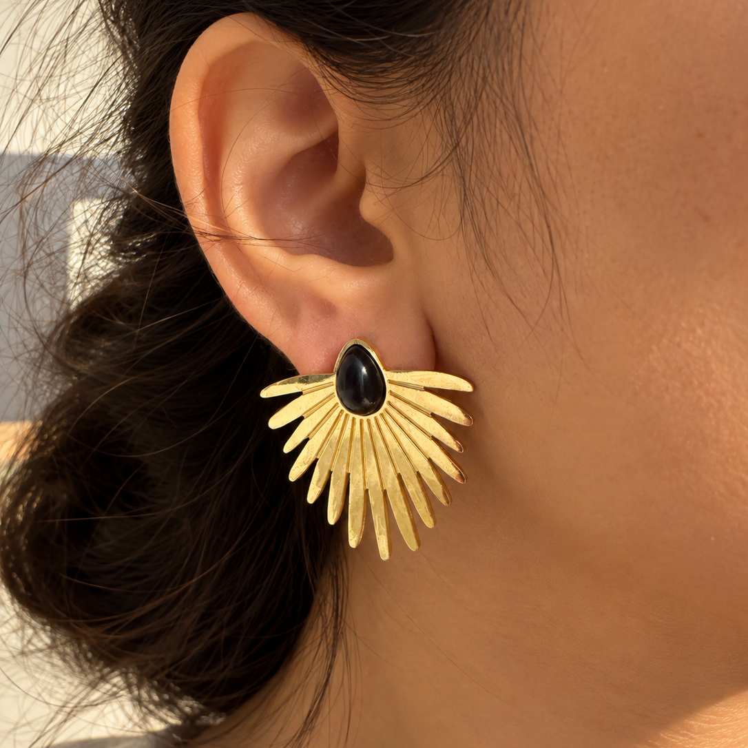 A close-up of a person's ear wearing a gold fan-shaped stud earring with a black teardrop-shaped stone at the center.