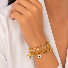 A close-up of a person's wrist wearing a stack of gold charms, including crescent moon, evil eye, and small bead charms, over a white top.