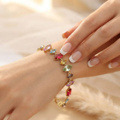 A gold-toned bracelet with multiple colorful gemstones, shown worn on a woman's wrist with her hand resting above it. The bracelet features marquise-cut stones in pink, blue, yellow, green, and purple on a gold chain.