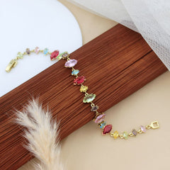 A gold-plated bracelet with multiple colorful marquise-cut gemstones in pink, green, yellow, blue, and purple, styled on a wooden block with a white plate and tulle fabric.