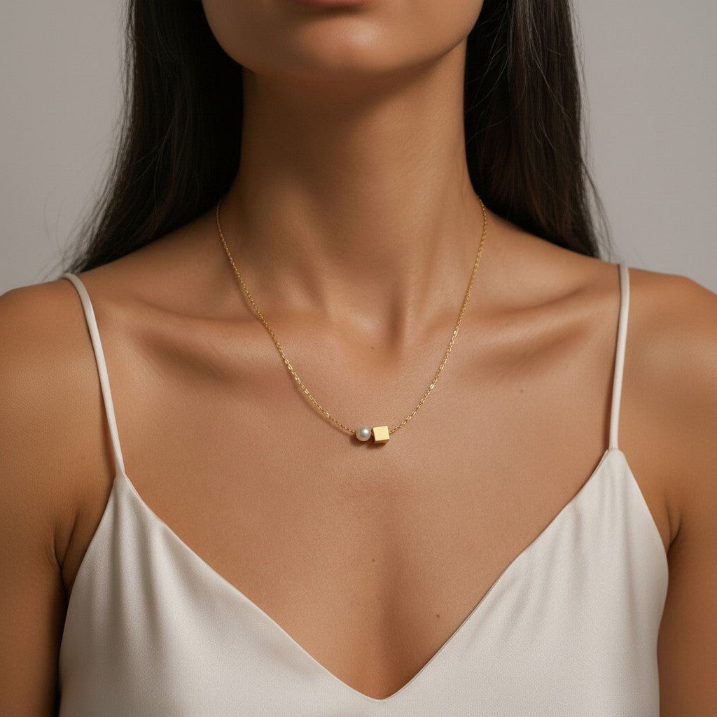 A close-up of a model wearing a delicate gold chain necklace with a small rectangular gold and iridescent pendant, styled with a white camisole against a neutral gray background.