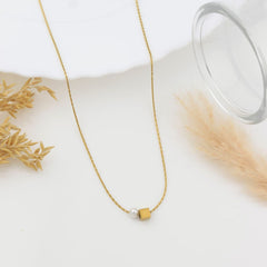 A delicate gold chain necklace with a small gold cube pendant and a single white pearl bead, styled on a white surface with dried grass and a glass prop.