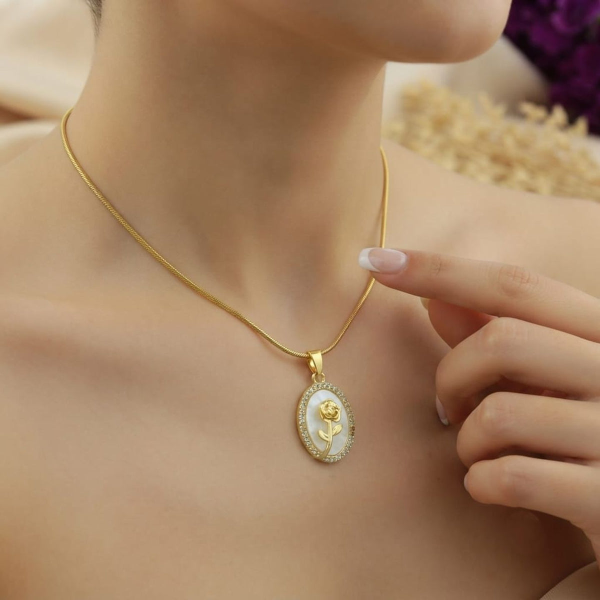 A close-up lifestyle shot of a woman wearing a gold snake chain necklace with an oval pendant. The pendant features a white background with a gold rose design and a border of small clear stones.