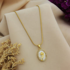 A gold chain necklace with an oval pendant featuring a white background and a gold rose design, displayed on a beige fabric backdrop with dried flowers.