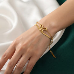A delicate gold double-strand bracelet with a bow-shaped charm and a small dangling tag, shown worn on a person's wrist against a white and dark green background.