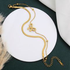 A gold snake chain bracelet with a central bow-shaped charm, displayed on a white circular stand against a dark green background with soft decorative props.