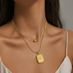 A close-up of a model's neck and upper chest wearing a double-layered gold necklace. The top layer is a fine chain with a small gold bead, and the bottom layer features a polished gold rectangular pendant with a tiny clear gemstone in the center.