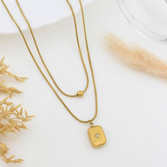 A gold-toned double-strand necklace with a small rectangular pendant featuring a star detail, styled on a white surface with dried grass accents.
