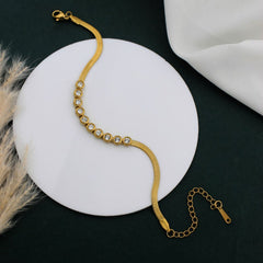 A gold snake-chain bracelet with a row of clear rhinestones, displayed on a round white pedestal against a dark background with decorative dried grass and white fabric.