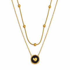A double-layered gold necklace featuring a longer chain with spaced gold beads and a shorter chain with a round black pendant containing a gold heart, shown on a plain white background.