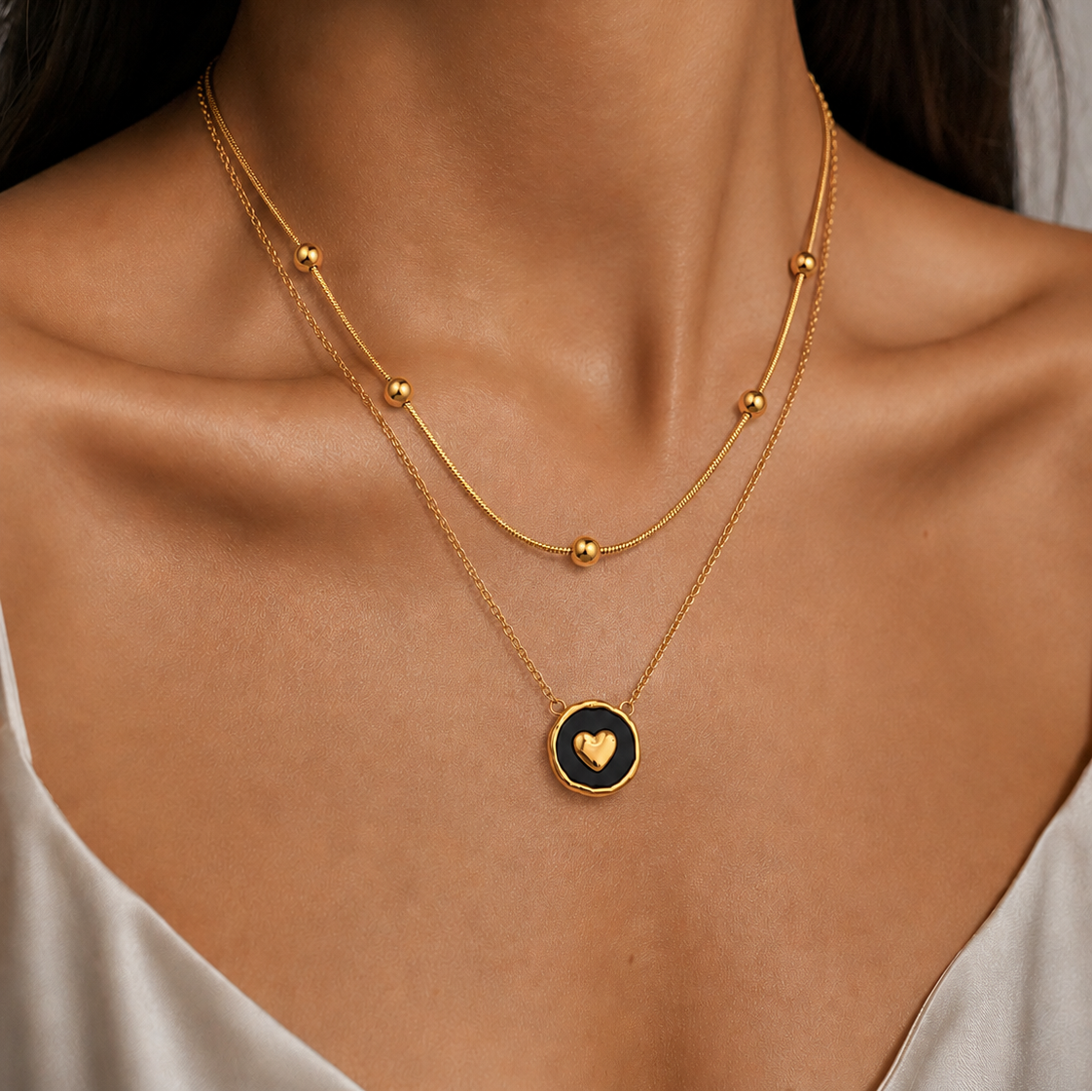 A close-up of a model's neck and chest wearing a double-layered gold necklace. The necklace features a delicate chain with small gold beads and a circular pendant with a black center and a gold heart.