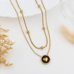 A double-strand gold necklace with a snake chain and beaded chain, featuring a round black pendant with a gold heart, displayed on a white surface with dried botanical props.