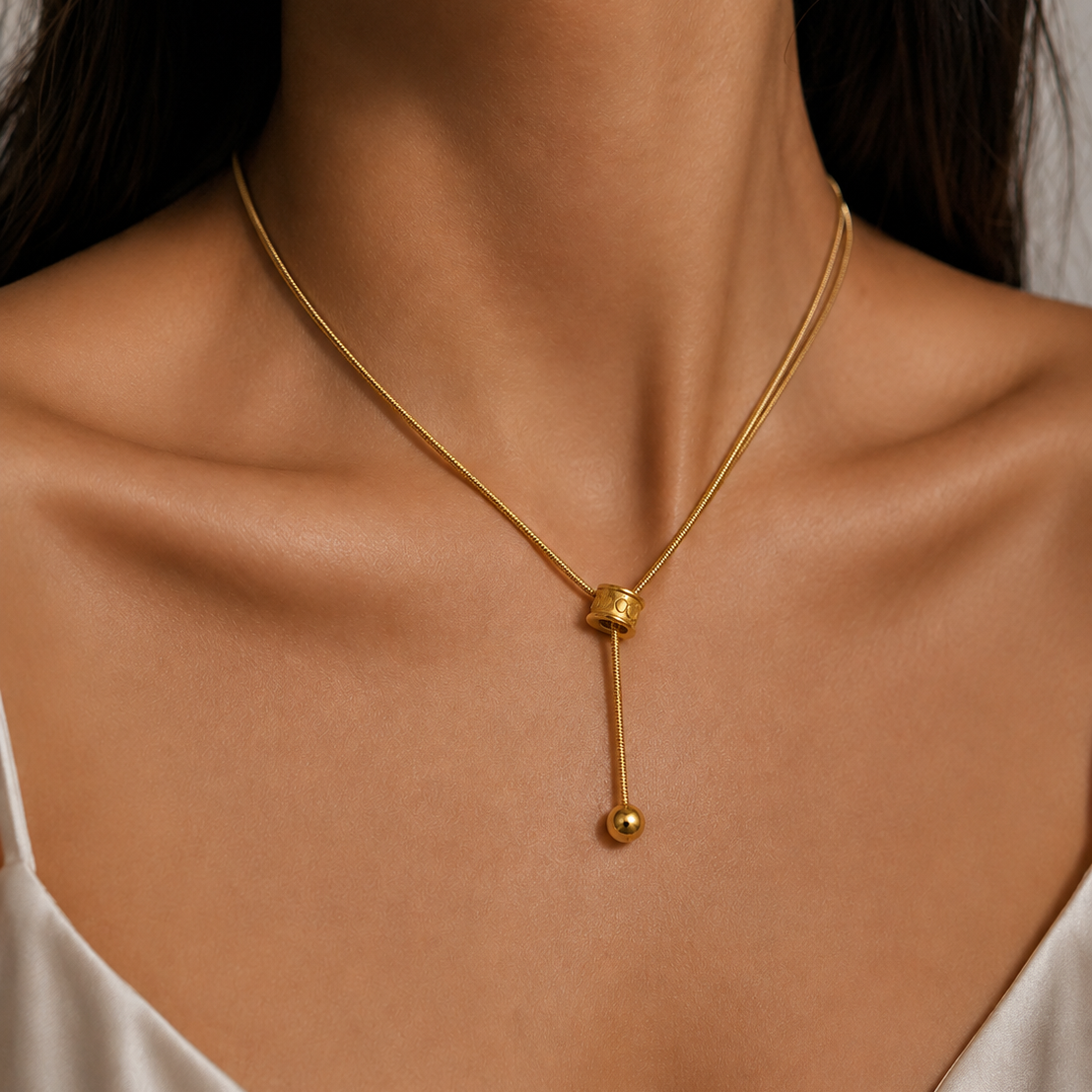 A close-up of a model's neck and upper chest wearing a delicate gold chain necklace with a small spherical charm and a dangling bead.