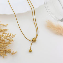 A delicate gold necklace with a double-strand chain, a textured cylindrical bead, and a small spherical bead pendant, styled on a white surface with dried grass and a glass rim.