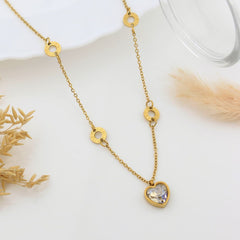 A gold chain necklace with three small circular gold charms and a heart-shaped pendant containing a clear faceted gemstone, styled on a white surface with dried grass and a glass rim.