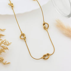 A gold-toned chain necklace with three knotted heart-shaped charms, styled on a white surface with dried grass and a glass rim in the background.