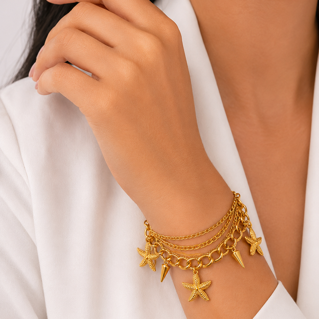 A close-up of a model's wrist wearing a delicate gold multi-layer bracelet with starfish and conical charms, styled with a white blazer.