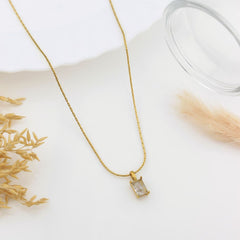 A delicate gold chain necklace with a small rectangular clear gemstone pendant, styled on a white surface with dried grass and a glass prop.