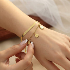 A close-up of a person's wrist wearing two delicate gold bracelets: a textured snake-style chain bracelet and a thinner chain bracelet with small star charms, with a hand gently touching the lower charm.