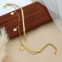 A gold-toned double-layer necklace with a snake chain and delicate chain links adorned with starburst charms, styled on a wooden block over a beige surface with soft decorative props.
