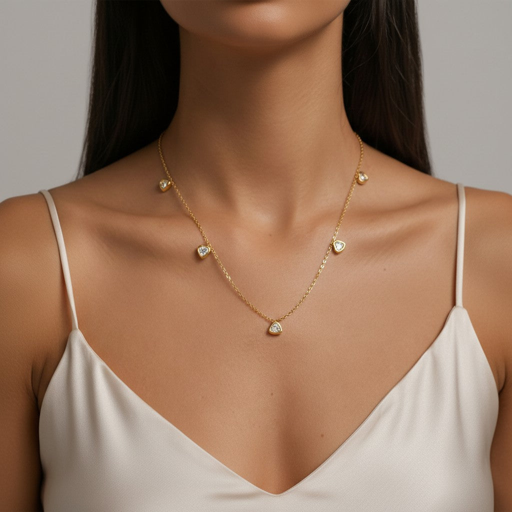 A close-up of a model wearing a delicate gold chain necklace with small heart-shaped crystal pendants, styled with a cream-colored spaghetti strap top against a neutral gray background.
