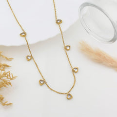 A gold chain necklace with seven small heart-shaped clear stones spaced along the chain, styled on a white surface with dried grass and a glass bowl prop.