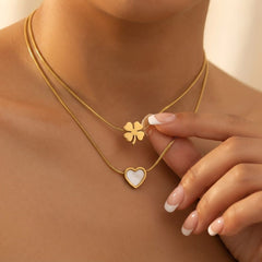 A close-up of a model wearing a double-layered gold necklace. The top layer features a small gold four-leaf clover pendant, while the bottom layer has a gold heart pendant with a white inlay.